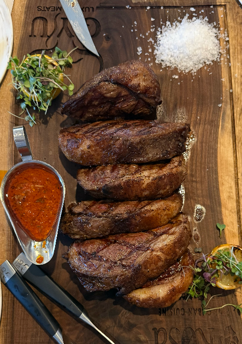 Grilled steaks on a wooden cutting board with a side of sauce and herbs.